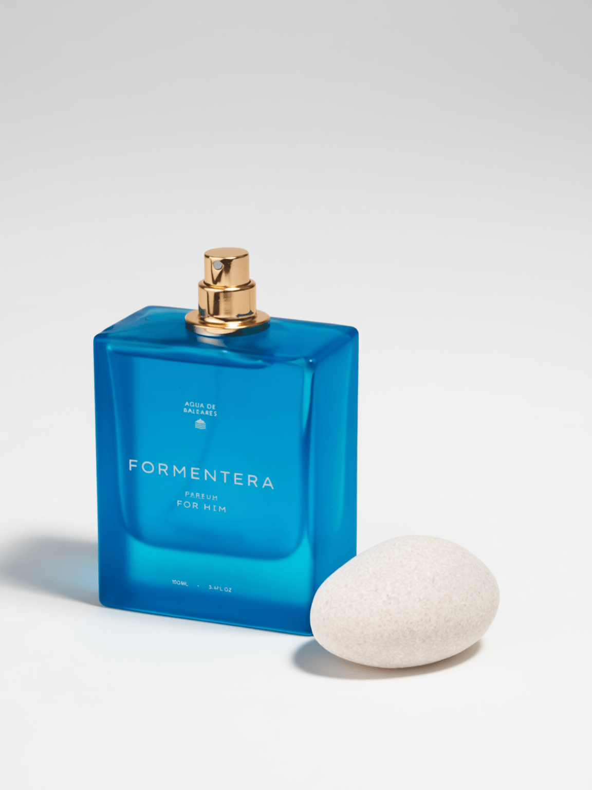 A blue rectangular Formentera for Him perfume bottle with a gold spray top, shown next to a smooth white stone on a white background.