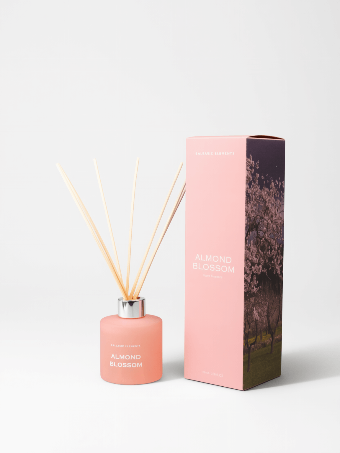A pink reed diffuser bottle with natural reed sticks next to its pink packaging box featuring a cherry blossom tree design, on a light gray background.