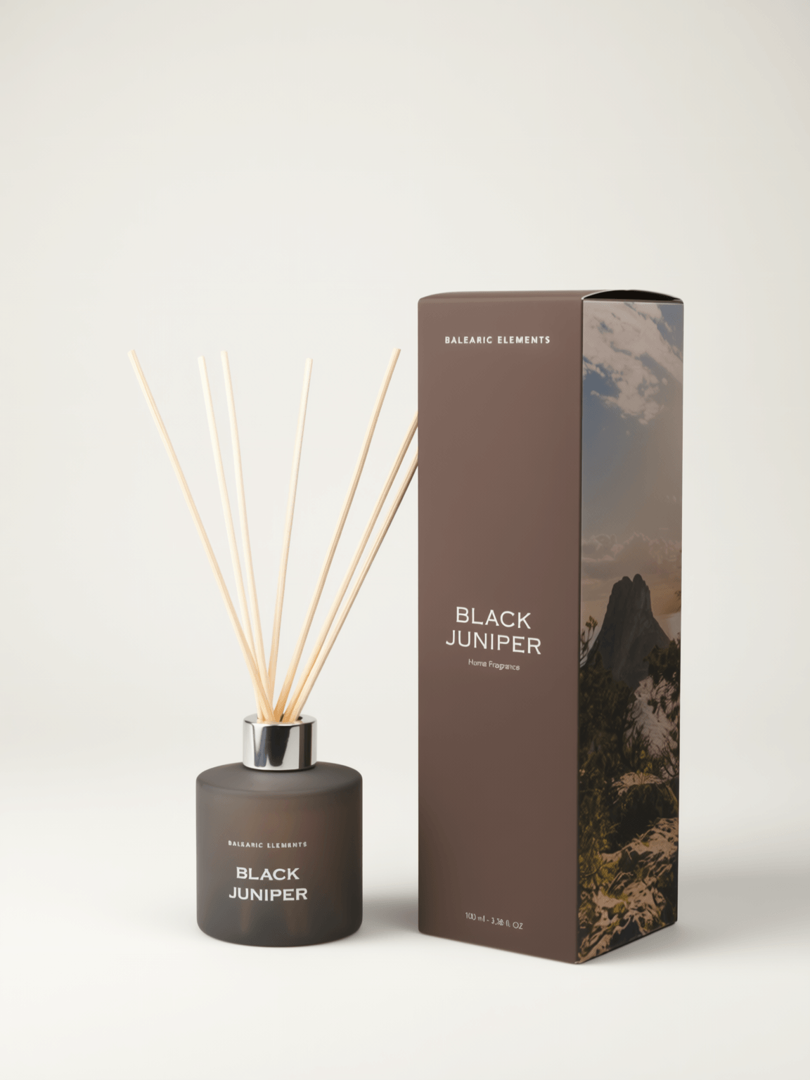 A reed diffuser bottle with light natural reeds next to its tall branded box on a clean off-white background. The bottle is dark charcoal gray with white text reading BLACK JUNIPER, and the box is brown with a mountain landscape print and the same product name.