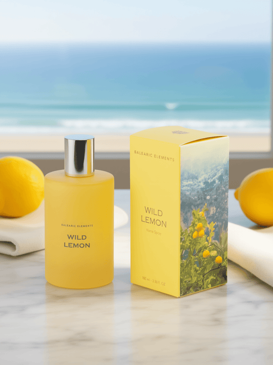 A yellow Balearic Elements Wild Lemon fragrance bottle stands next to its matching yellow box on a marble surface, with lemons and a blurred ocean view in the background.