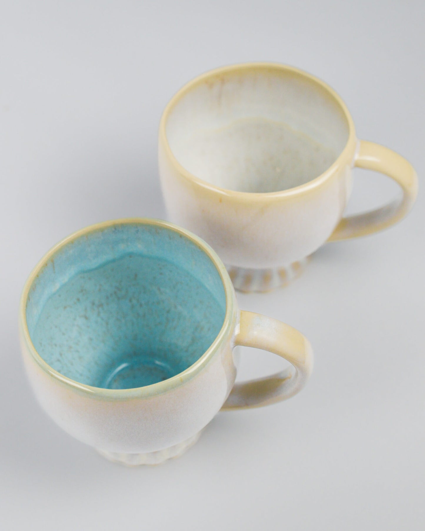 Two large ceramic mugs, one with a blue interior and cream exterior, the other with an off-white interior and cream exterior, on a white background.