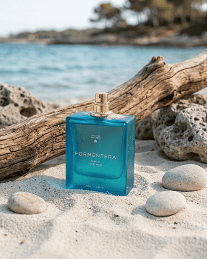 A blue rectangular perfume bottle labeled FORMENTERA PARFUM FOR HER with a gold spray top, placed on white sand with driftwood and rocks, with a blurred beach and ocean background.