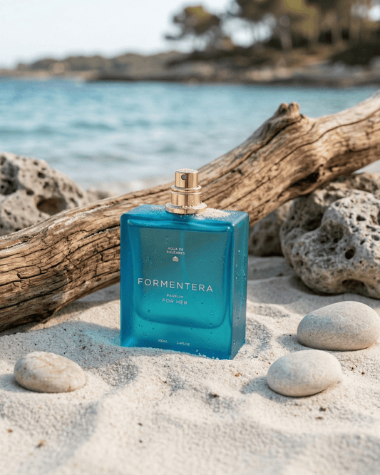 A blue rectangular perfume bottle labeled FORMENTERA PARFUM FOR HER with a gold spray top, placed on white sand with driftwood and rocks, with a blurred beach and ocean background.