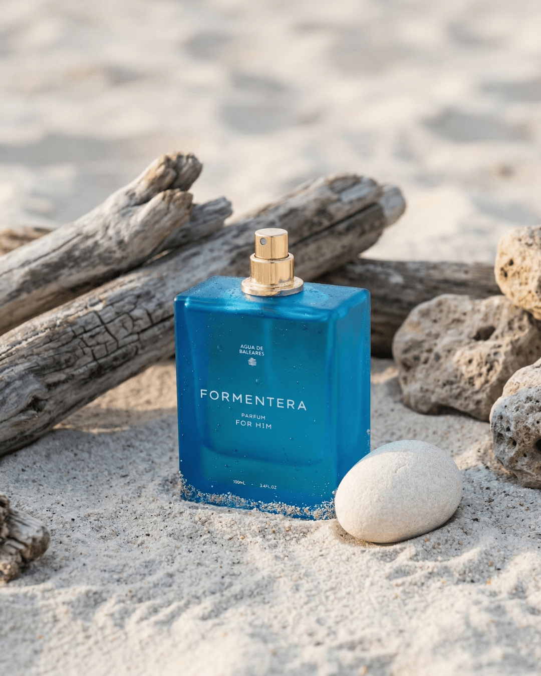 A blue rectangular perfume bottle labeled FORMENTERA stands on sandy ground with driftwood logs and rocks around it, creating a beach-like setting.