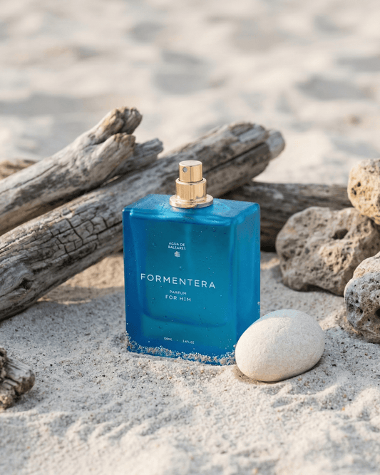 A blue rectangular perfume bottle labeled FORMENTERA stands on sandy ground with driftwood logs and rocks around it, creating a beach-like setting.