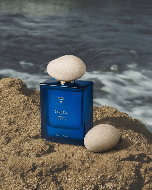 A blue rectangular perfume bottle labeled 'IBIZA' sits on a sandy rock with two smooth stones balancing it, with ocean waves blurred in the background.