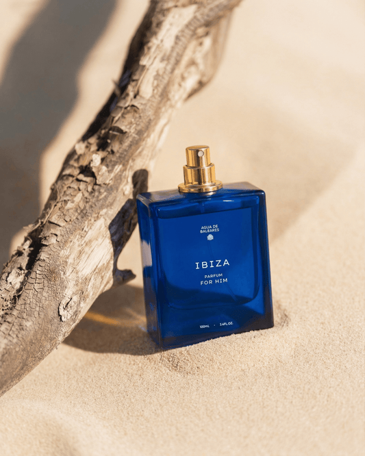 A blue rectangular perfume bottle labeled IBIZA PARFUM FOR HIM with a gold spray top, placed on sand next to a piece of driftwood.