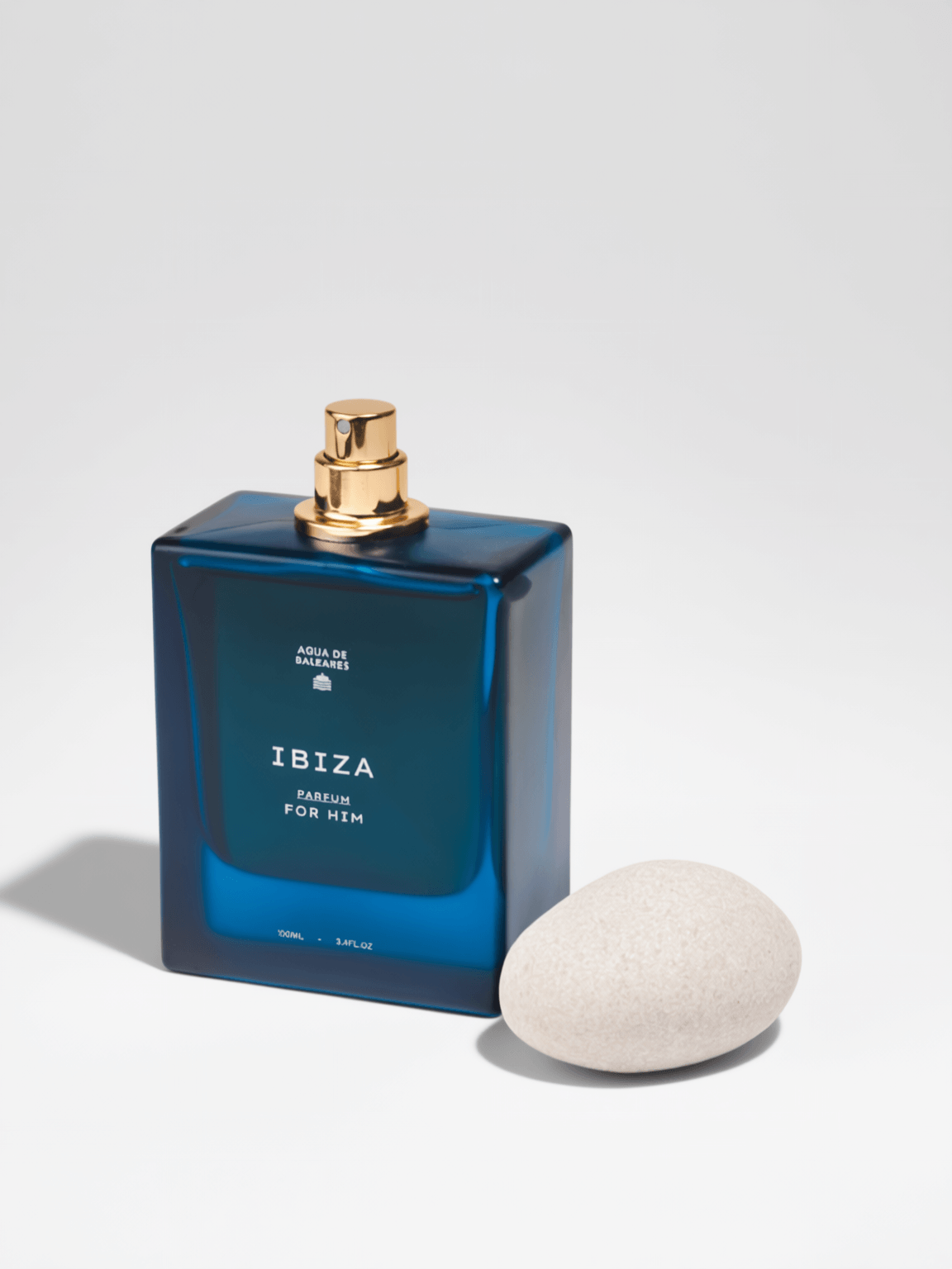 A rectangular blue glass perfume bottle labeled 'IBIZA PARFUM FOR HIM' with a gold spray top, shown next to a smooth light beige stone on a clean white background.