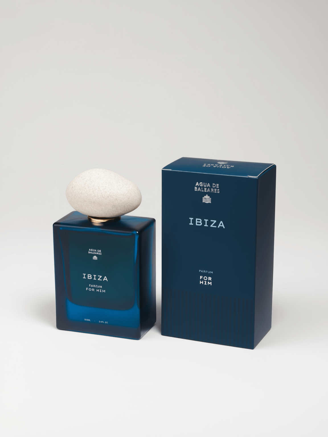 A dark blue rectangular perfume bottle with a white rounded stone-like cap stands beside its matching dark blue box on a white studio background. The bottle and box are labeled IBIZA 