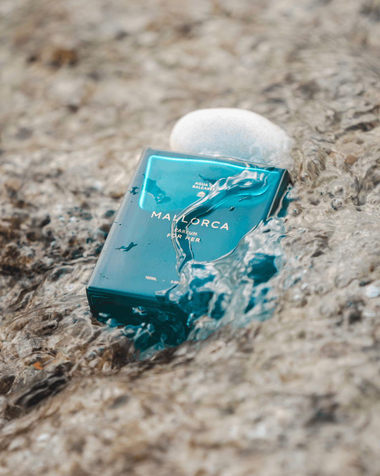 A teal rectangular perfume bottle labeled 'MALLORCA' sits in shallow water on a sandy beach, with a white stone resting on top.