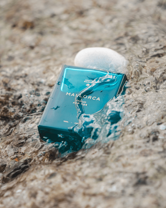 A teal rectangular perfume bottle labeled 'MALLORCA' sits in shallow water on a sandy beach, with a white stone resting on top.