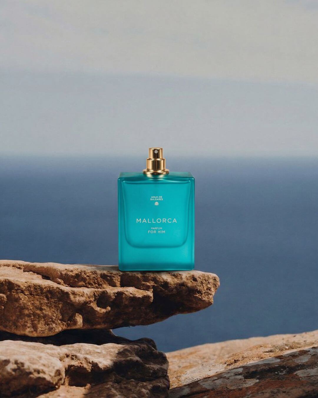 A turquoise rectangular perfume bottle with a gold spray top stands on a rocky outcrop with a blurred ocean and sky background. The bottle is labeled Mallorca.