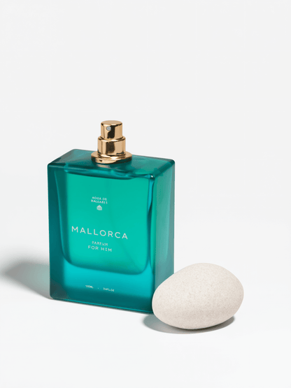 Mallorca Eau de Parfum for men in a translucent teal-green rectangular glass bottle with a gold spray cap, shown next to a smooth white stone on a white background.