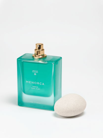 A rectangular translucent aqua-green perfume bottle labeled MENORCA with a gold spray top, photographed on a clean white background with a smooth off-white stone beside it.
