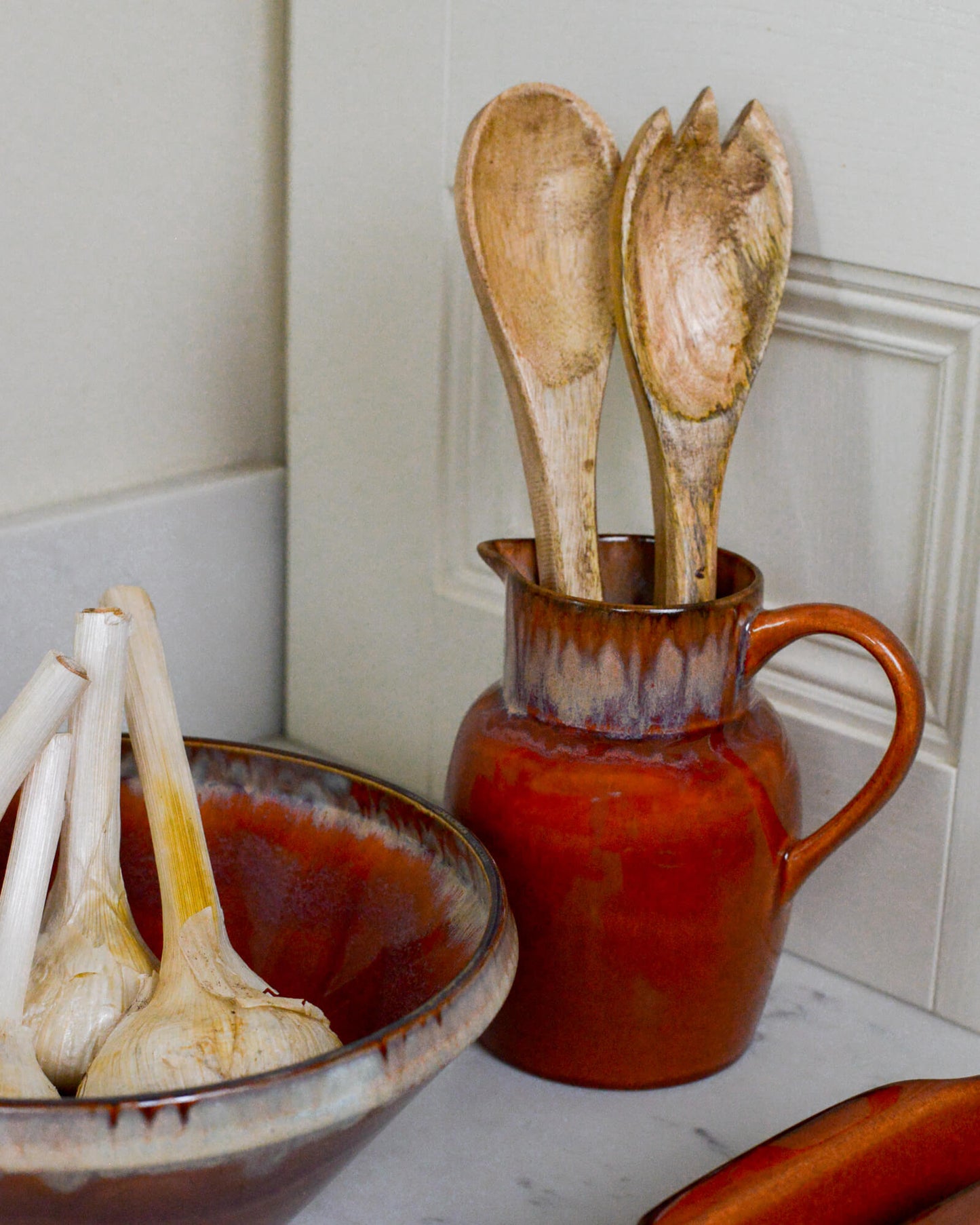 Burnt orange glazed ceramic pitcher with wooden utensils against a white tiled wall.