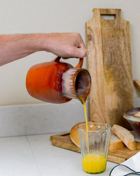 Person pouring orange juice from a ceramic pitcher into a glass on a kitchen counter.