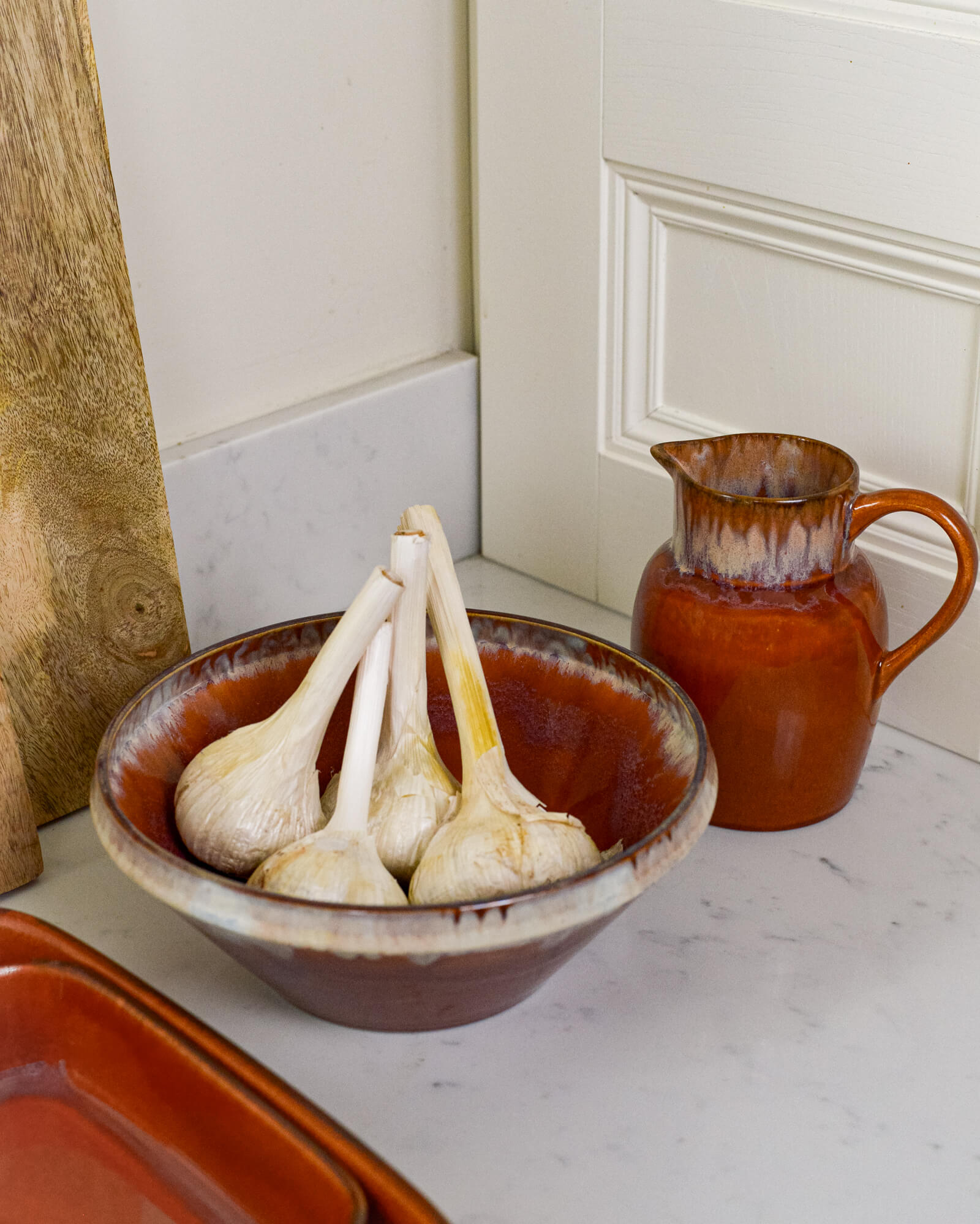 Burnt orange ceramic bowl with garlic and matching pitcher on a white marble surface
