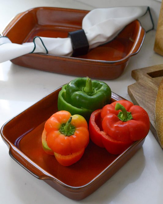 Three bell peppers (red, green, and orange) on a caramel square ceramic baking dish with a white linen napkin in the background.