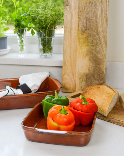 Three bell peppers (red, green, and orange) on a caramel square ceramic baking dish with a wooden chopping board and a white linen napkin in the background.