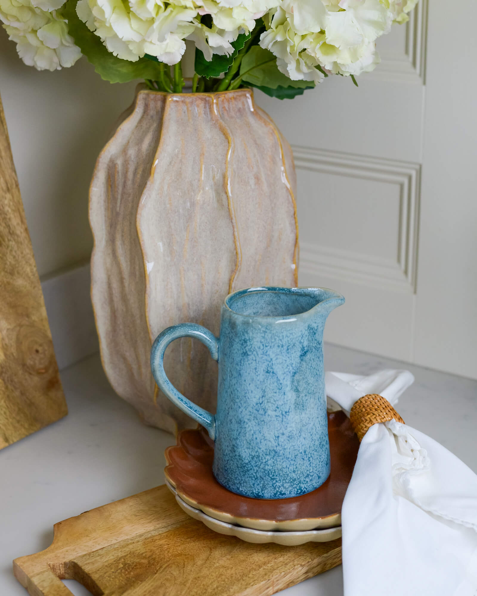 Blue ceramic pitcher on a wooden tray with a beige vase and flowers in the background.