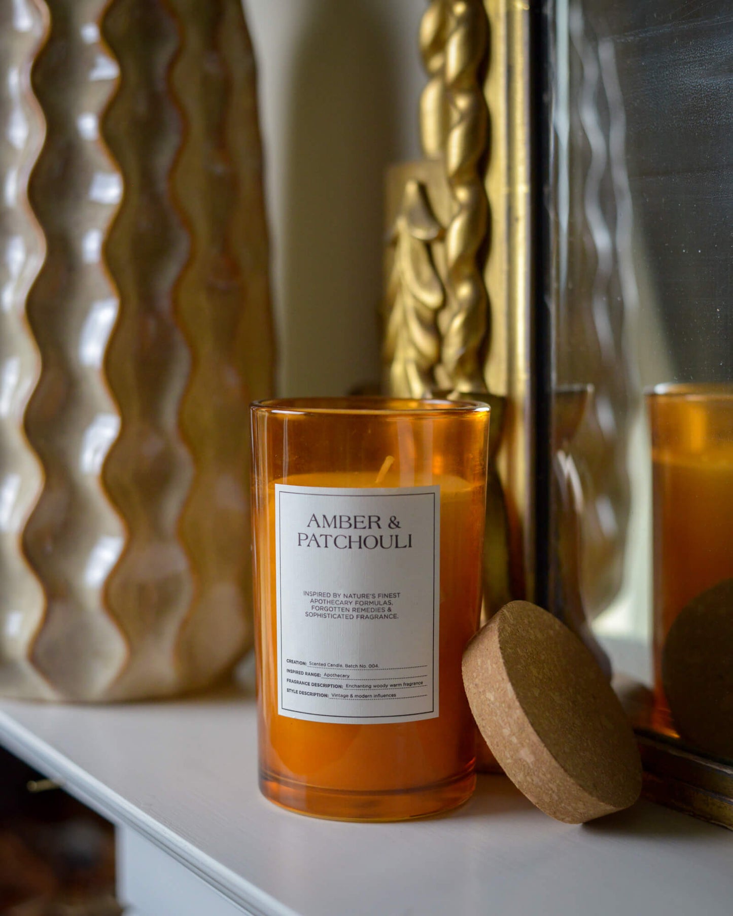 Amber-colored candle labeled 'Amber & Patchouli' on a decorative surface with a gold mirror and textured wall in the background.