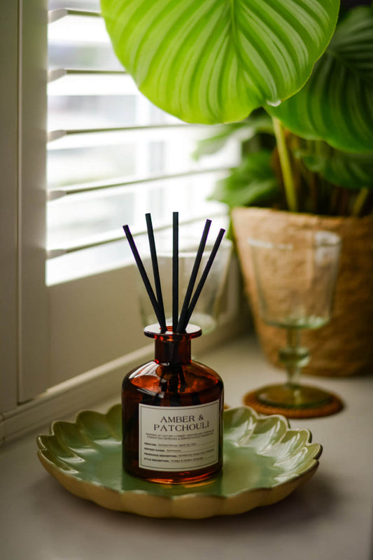 Reed diffuser labeled 'Amber & Patchouli' on a green scalloped edge tray with a plant in the background.