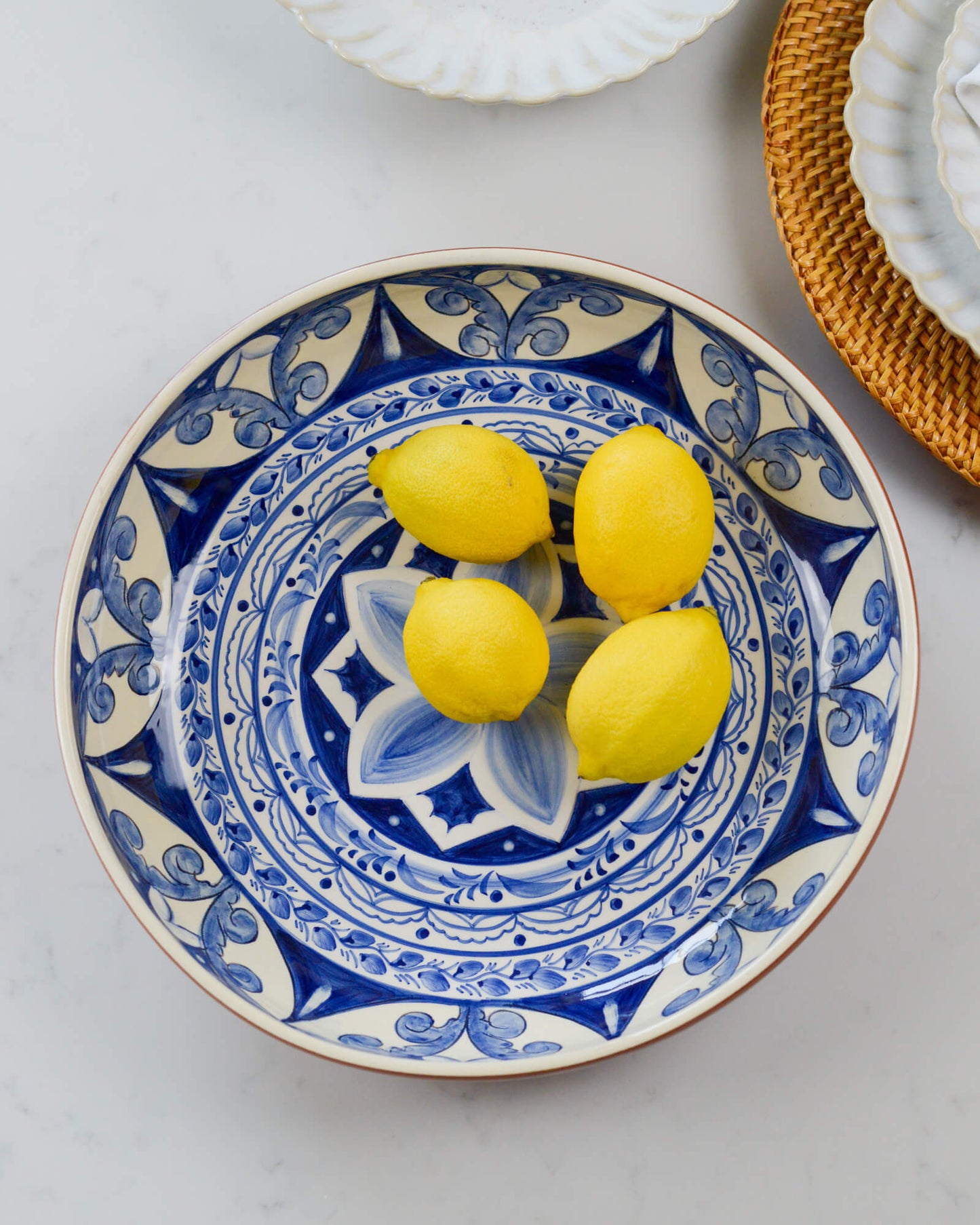 Decorative terracotta serving bowl with blue patterns holding three lemons on a light marble background