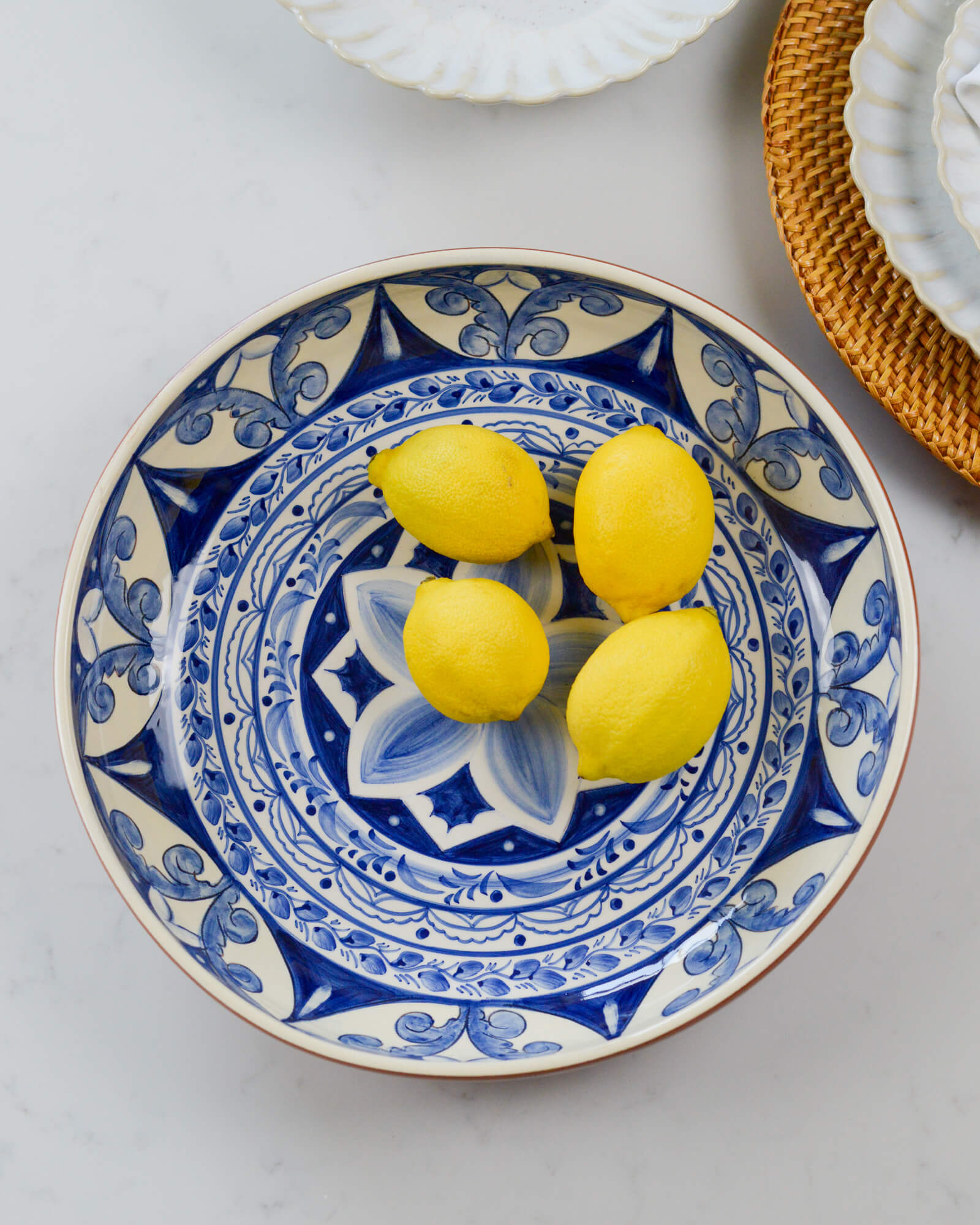 Decorative terracotta serving bowl with blue patterns holding three lemons on a light marble background