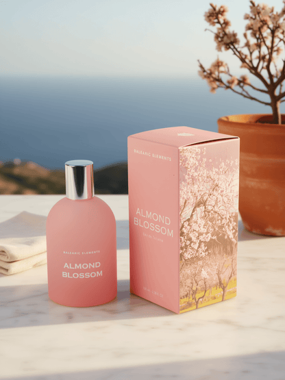 A pink perfume bottle labeled 'ALMOND BLOSSOM' stands beside its matching pink box with floral blossom artwork, placed on a light marble surface with a blurred outdoor coastal background and a potted plant.