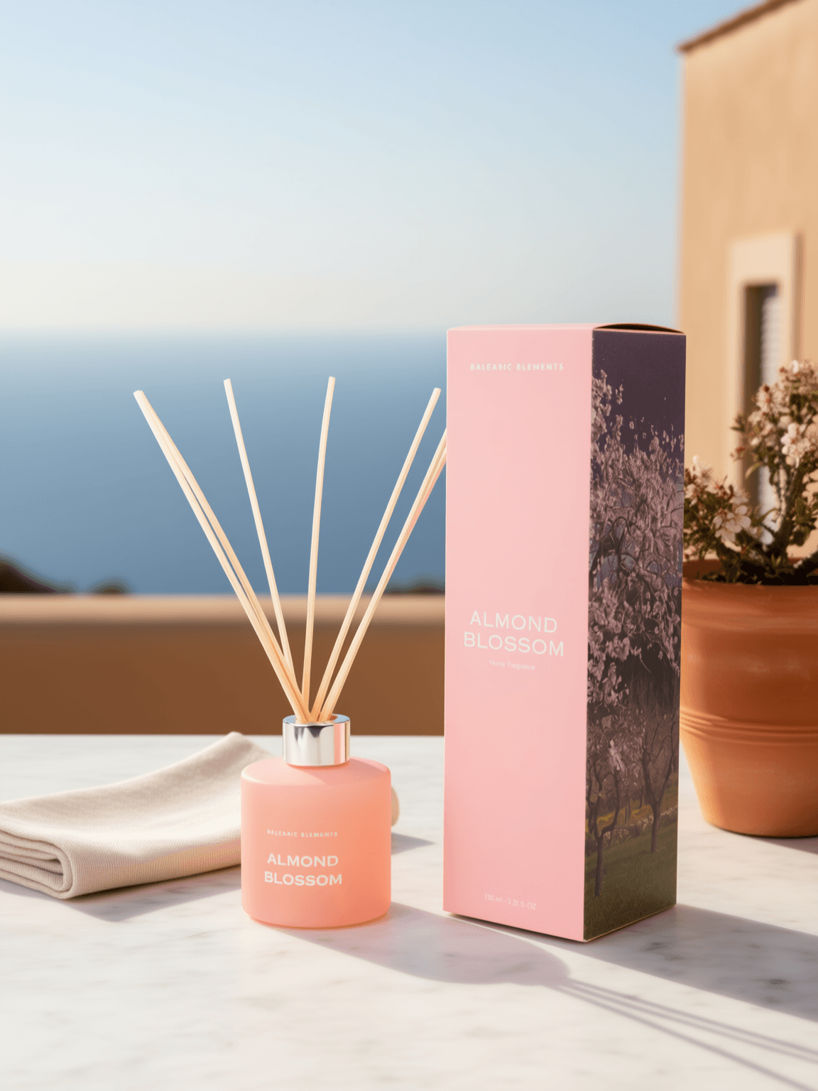 A pink reed diffuser bottle with natural reed sticks stands beside its matching pink box on a light stone surface outdoors. The box is labeled ALMOND BLOSSOM, and the bottle also shows the same fragrance name.