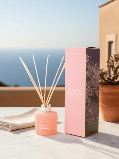 A pink reed diffuser bottle with natural reed sticks stands beside its matching pink box on a light stone surface outdoors. The box is labeled ALMOND BLOSSOM, and the bottle also shows the same fragrance name.