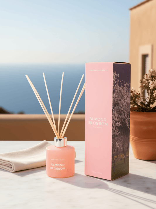 A pink reed diffuser bottle with natural reed sticks stands beside its matching pink box on a light stone surface outdoors. The box is labeled ALMOND BLOSSOM, and the bottle also shows the same fragrance name.