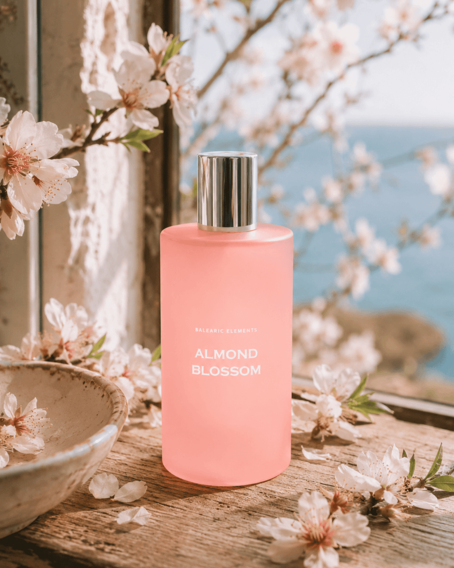 A pink bottle of Almond Blossom Room Spray stands on a rustic wooden surface surrounded by white blossoms, with a soft-focus outdoor background of blue water and sky.