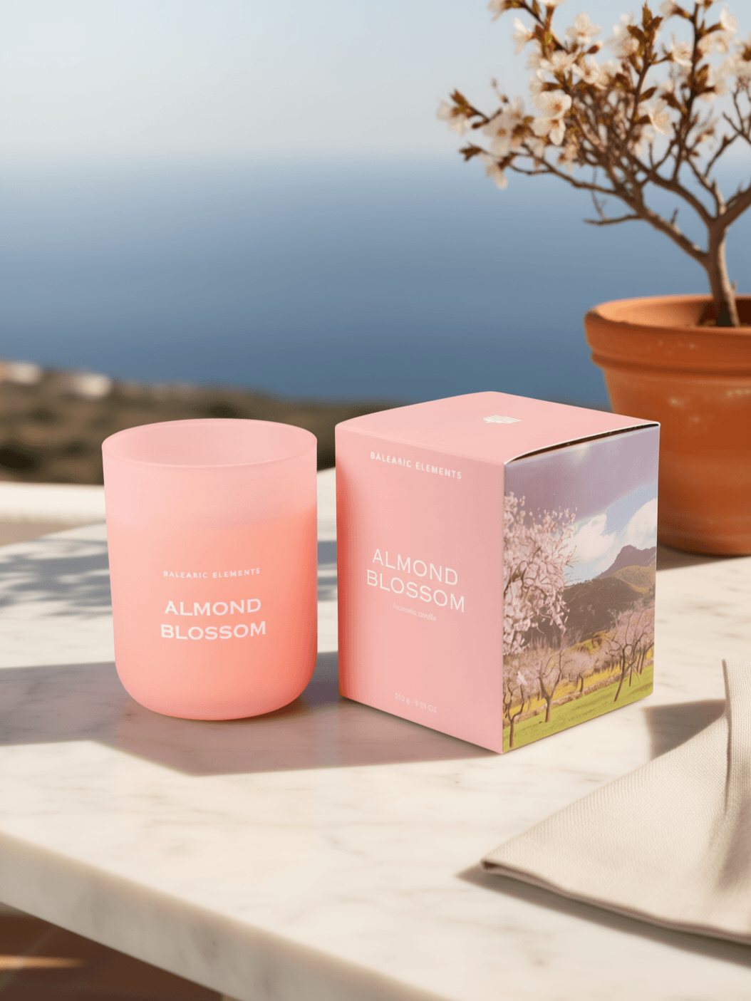 A pink glass candle jar and its matching pink box labeled 'ALMOND BLOSSOM' sit on a light stone surface outdoors, with a blurred coastal background and a potted flowering tree behind them.