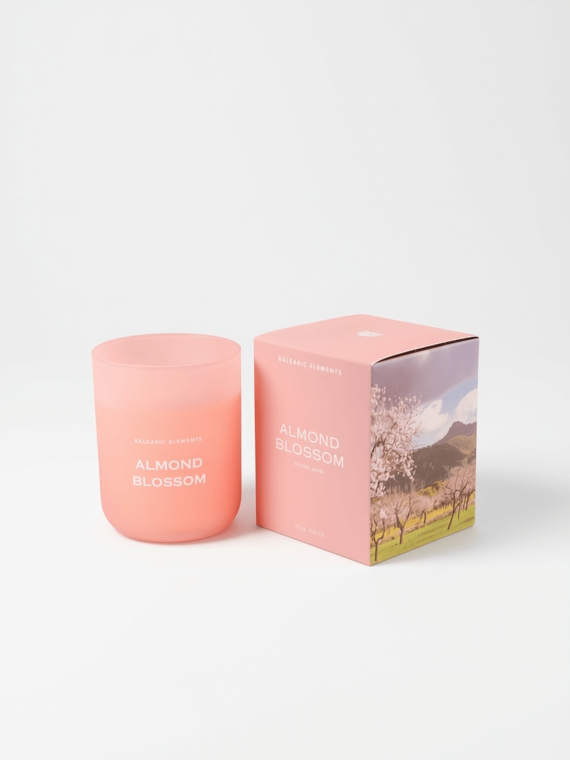 A pink glass jar candle labeled 'ALMOND BLOSSOM' stands next to its matching pink packaging box on a clean white background. The box features a scenic print of a blossoming tree and mountains.