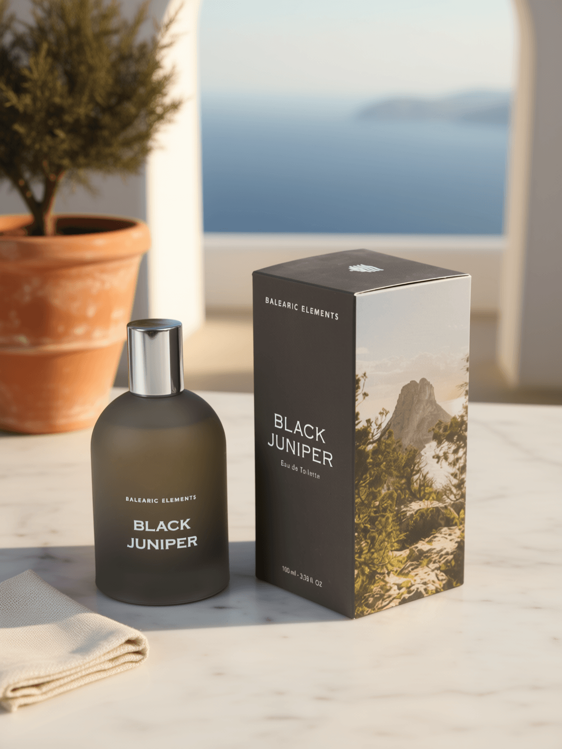 A bottle of Balearic Elements Black Juniper Eau de Toilette next to its packaging box, styled on a marble surface with a potted plant and a scenic ocean background.