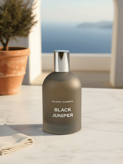 A matte dark gray bottle labeled 'BALEARIC ELEMENTS BLACK JUNIPER' with a reflective silver cap, set on a light marble surface with a blurred potted plant and sea view in the background.