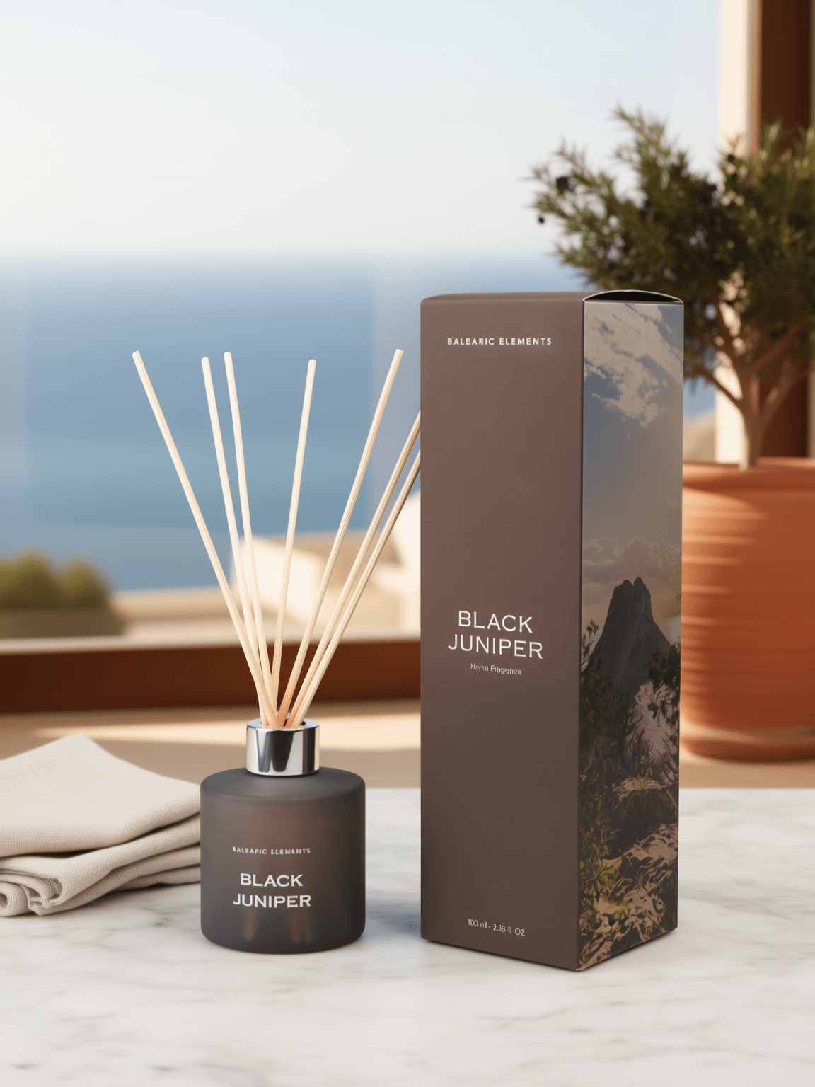 A reed diffuser bottle with light wooden reeds and its matching product box, labeled 'BALEARIC ELEMENTS BLACK JUNIPER', displayed on a marble surface with a blurred coastal background.