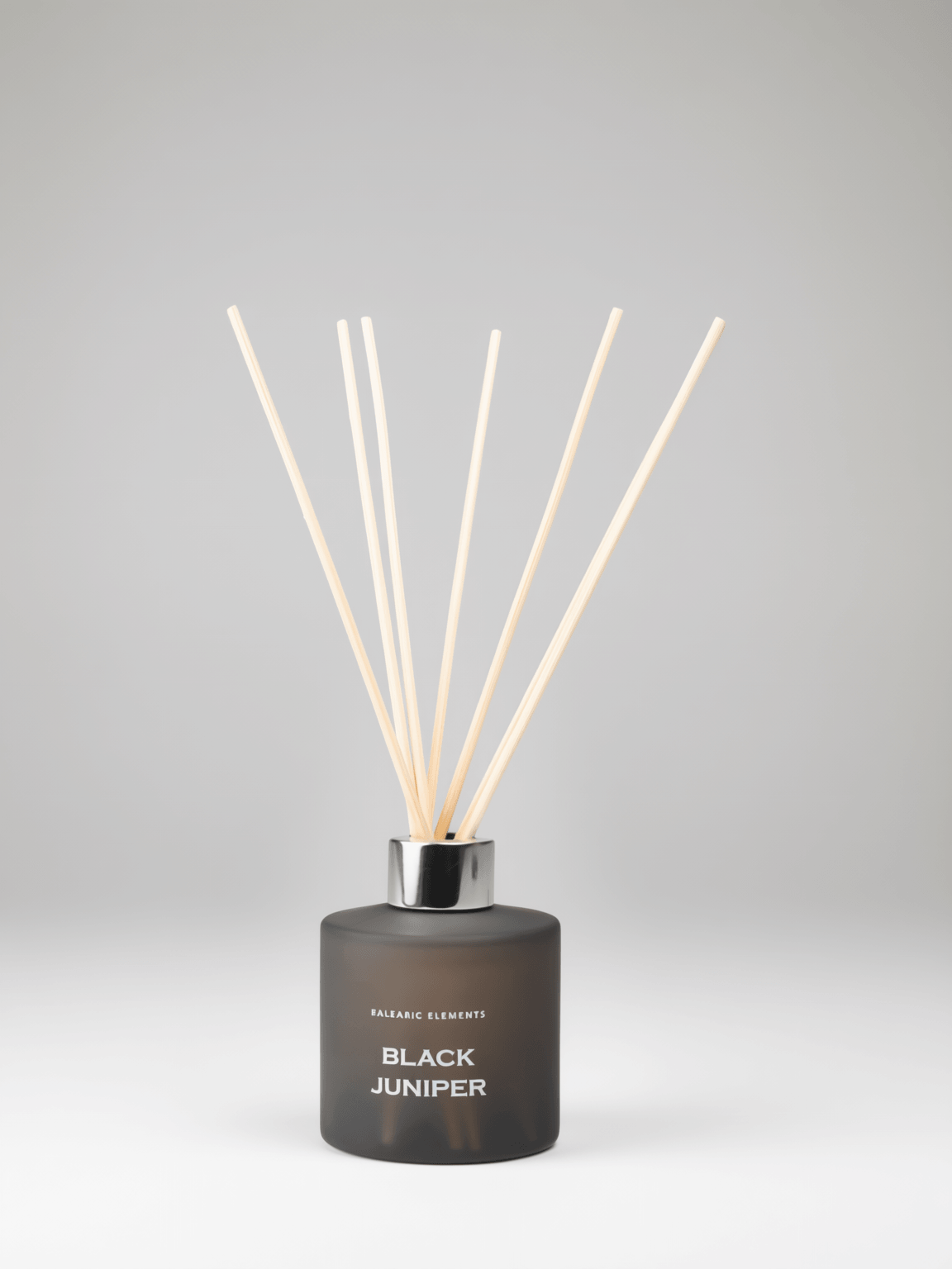 A reed diffuser bottle with a frosted dark gray glass body, a silver collar, and light natural wood reeds, labeled 'balearic ELEMENTS BLACK JUNIPER'.