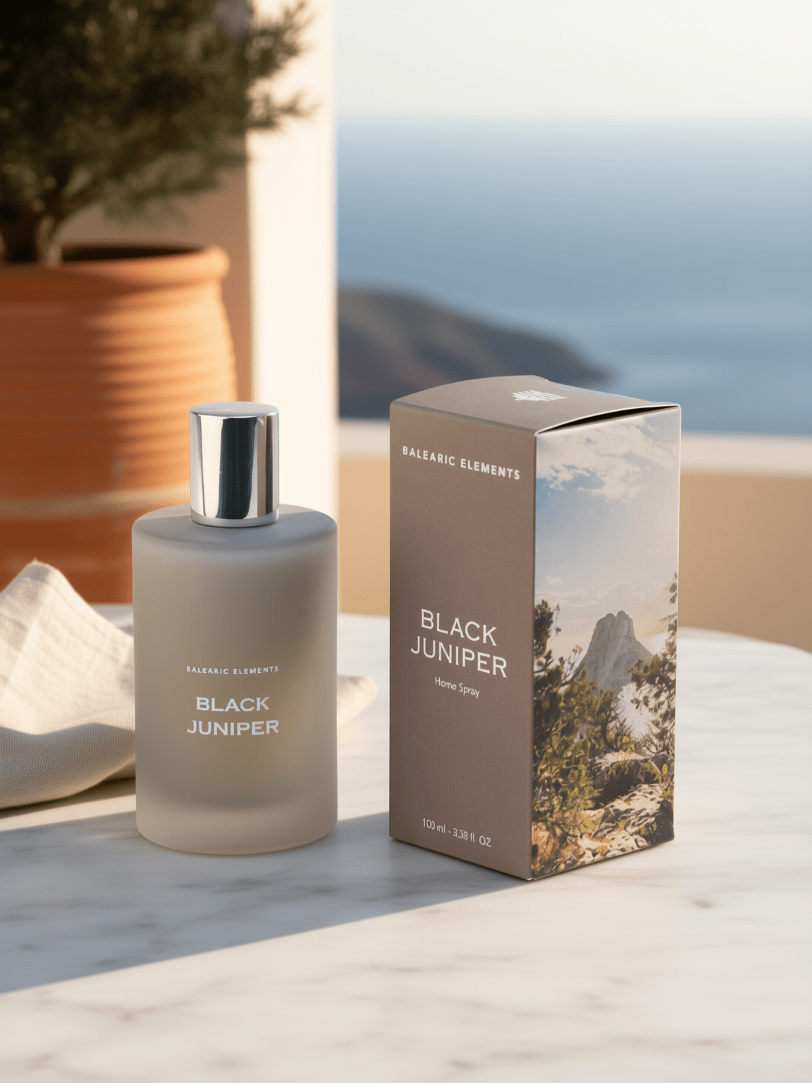 A frosted glass perfume bottle labeled 'BALEARIC ELEMENTS BLACK JUNIPER' stands beside its matching rectangular box on a light marble surface. The box features a scenic mountain landscape print, and the background shows a softly blurred outdoor coastal scene with warm sunlight and a potted plant.