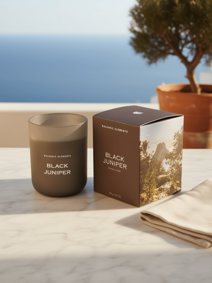 A matte dark brown glass jar candle labeled 'BLACK JUNIPER' stands beside its matching brown box on a light marble surface, with a folded cream cloth and a potted tree in the background against a blurred ocean view.
