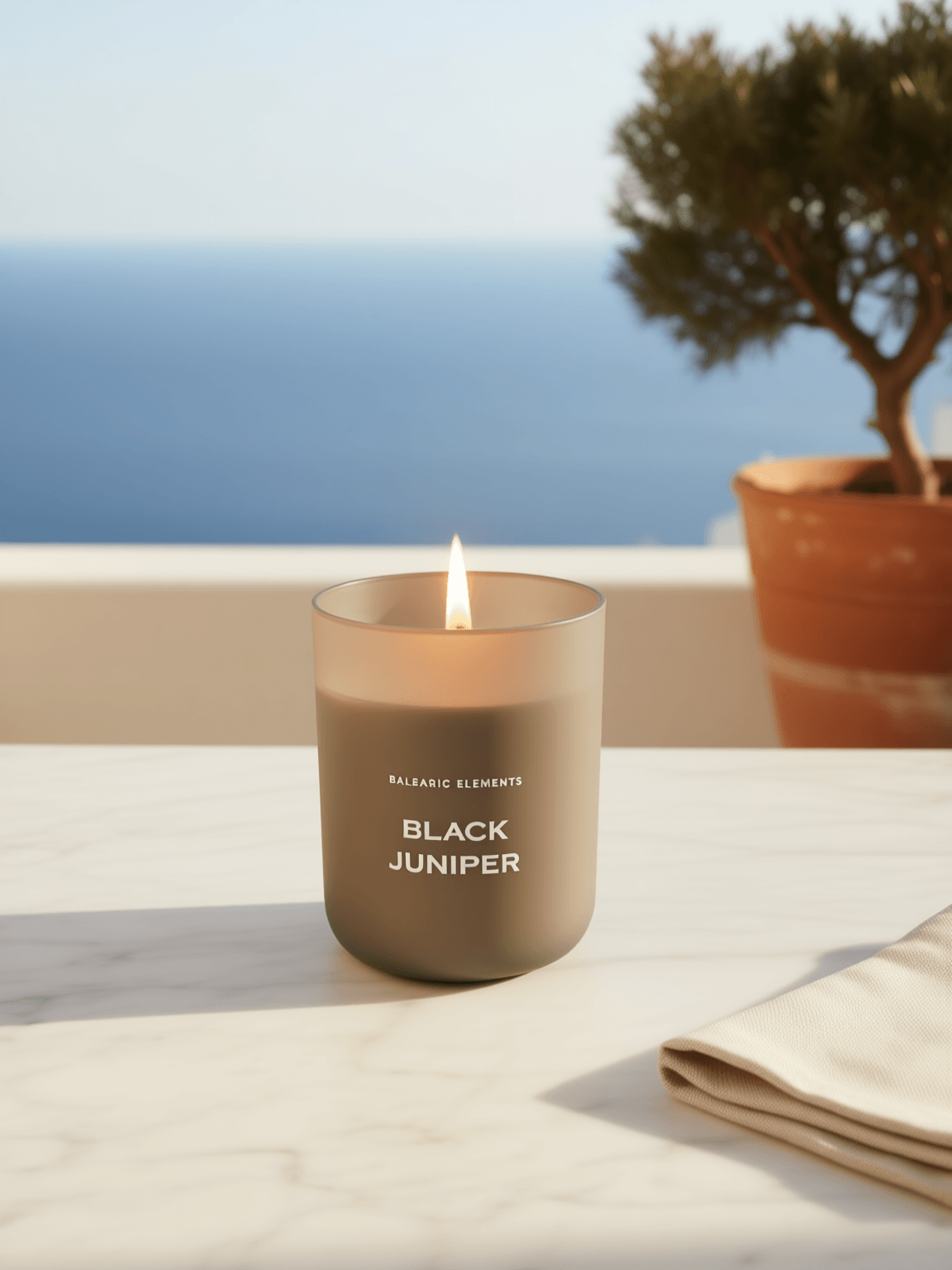 A lit matte taupe candle jar labeled 'BALEARIC ELEMENTS BLACK JUNIPER' sits on a white marble surface with an ocean and potted tree blurred in the background.
