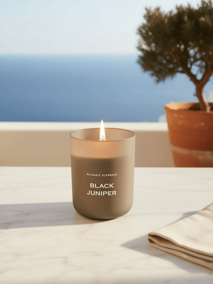 A lit matte taupe candle jar labeled 'BALEARIC ELEMENTS BLACK JUNIPER' sits on a white marble surface with an ocean and potted tree blurred in the background.