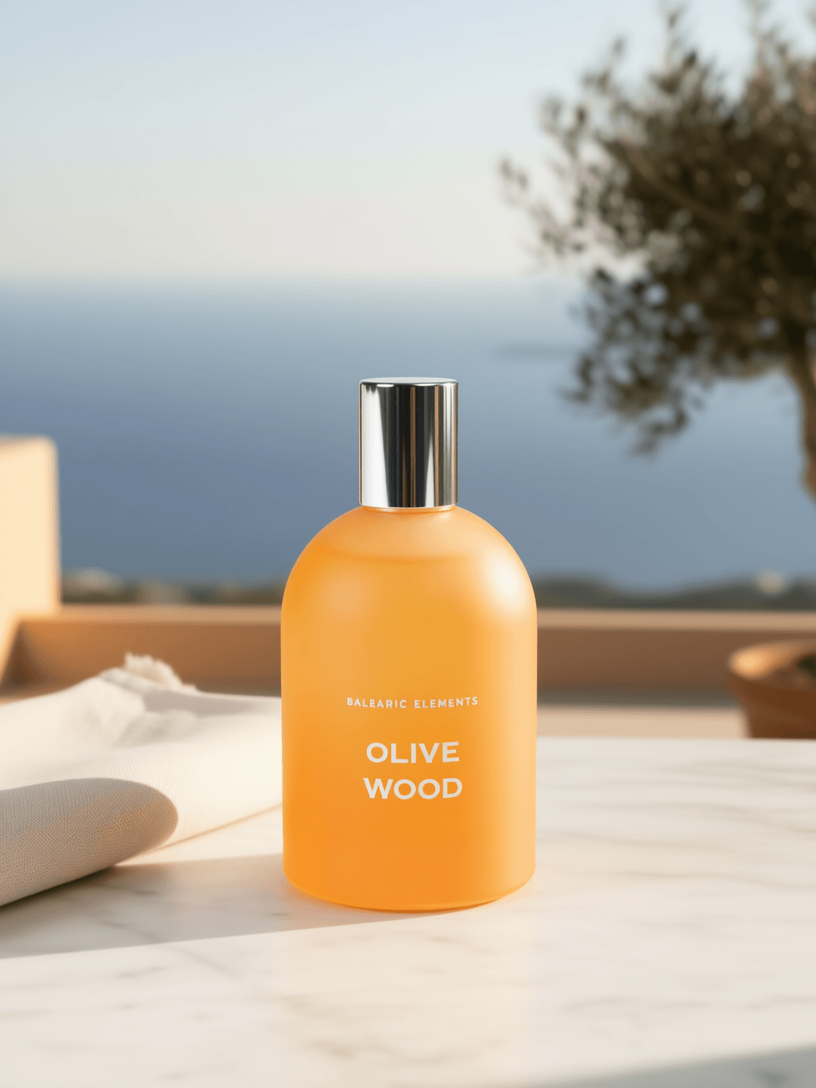 An orange bottle of Balearic Elements Olive Wood shower gel stands on a white marble surface with a blurred Mediterranean background showing the sea, sky, and a tree.