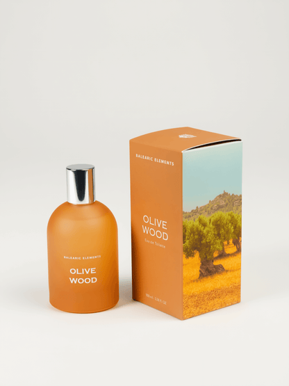 An orange frosted glass bottle with a shiny silver cap, next to it's box, labeled 'OLIVE WOOD' by Balearic Elements, set against a plain white background.