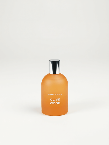 An orange frosted glass bottle with a shiny silver cap, labeled 'OLIVE WOOD' by Balearic Elements, set against a plain white background.