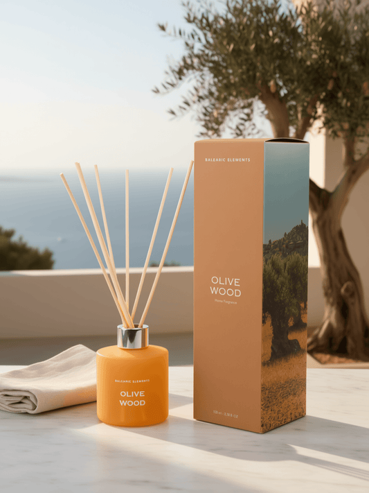 A reed diffuser bottle in amber orange with natural reed sticks, shown next to its matching tan and olive-green packaging box on a light stone surface, with a blurred outdoor background featuring a tree and sea.
