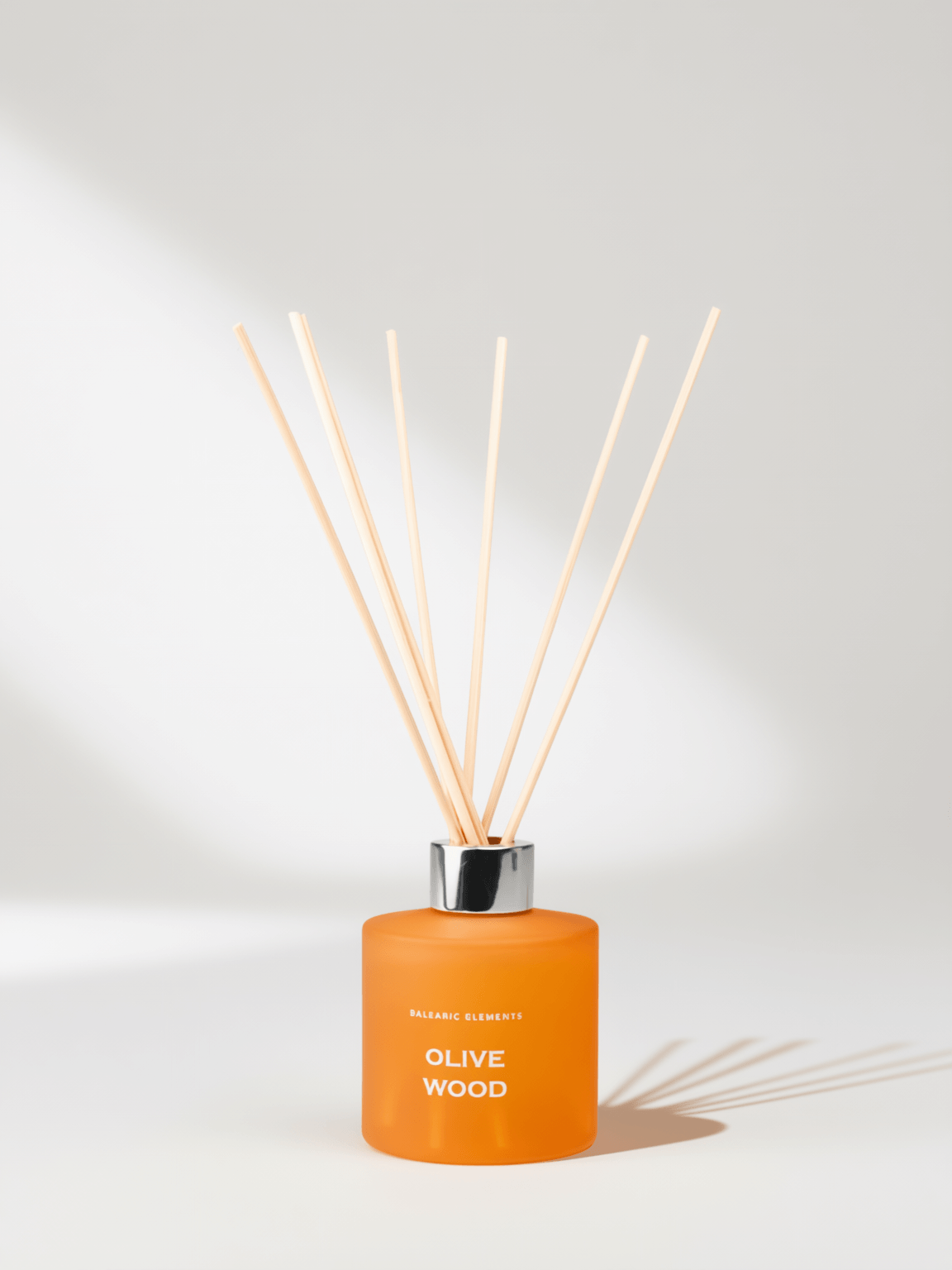 An orange reed diffuser bottle with a silver cap and natural wood reeds, labeled 'Olive Wood', on a light background with soft shadows.