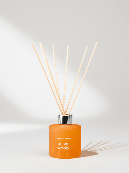 An orange reed diffuser bottle with a silver cap and natural wood reeds, labeled 'Olive Wood', on a light background with soft shadows.