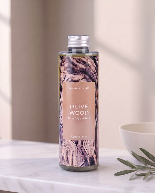 A bottle of Balancer Elements Olive Wood Home Fragrance Refill with a rustic wood grain label and silver cap, set on a marble countertop with a white bowl and olive leaves in a softly lit room.